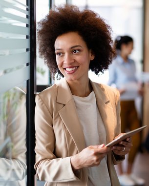 A woman with curly hair stands by a glass wall holding a tablet. She is wearing a light blazer and smiling. In the background, two other women can be seen talking to each other.