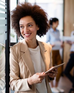 A woman with curly hair stands by a glass wall holding a tablet. She is wearing a light blazer and smiling. In the background, two other women can be seen talking to each other.