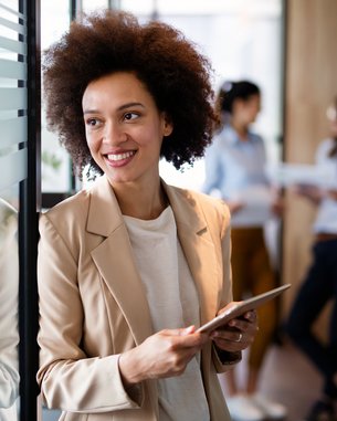 A woman with curly hair stands by a glass wall holding a tablet. She is wearing a light blazer and smiling. In the background, two other women can be seen talking to each other.