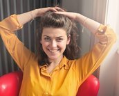 Eine junge Frau mit lockigem Haar sitzt in einem roten Stuhl. Sie trägt ein gelbes Hemd und lächelt, während sie ihre Hände hinter dem Kopf hält. Natürliches Licht fällt auf ihr Gesicht.