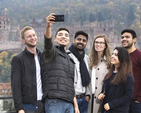 Sechs Personen machen ein Selfie vor einer historischen Burg und einer herbstlichen Landschaft.