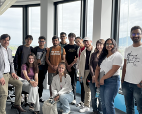 Gruppe von 16 Personen in einem modernen Büro mit großen Fenstern, einige sitzen, andere stehen, alle lächeln und posieren für ein Foto.