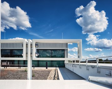 Moderne Architektur eines Gebäudes mit großen Fenstern und einem Flachdach, umgeben von einem klaren blauen Himmel mit weißen Wolken.