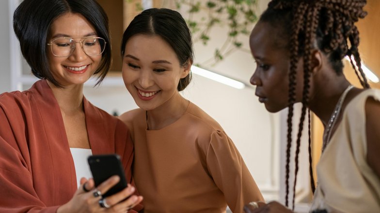 Drei Frauen stehen zusammen und schauen auf ein Smartphone. Eine Frau mit Brille lächelt, während die anderen beiden interessiert auf den Bildschirm blicken. Sie tragen modische, farblich abgestimmte Kleidung.