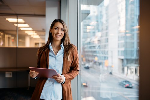 A smiling woman stands by a window in a modern office building, holding a tablet in her hand. She is wearing a brown jacket and a light blue shirt. In the background, city views and office furnishings are visible.