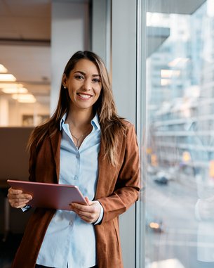 A smiling woman stands by a window in a modern office building, holding a tablet in her hand. She is wearing a brown jacket and a light blue shirt. In the background, city views and office furnishings are visible.
