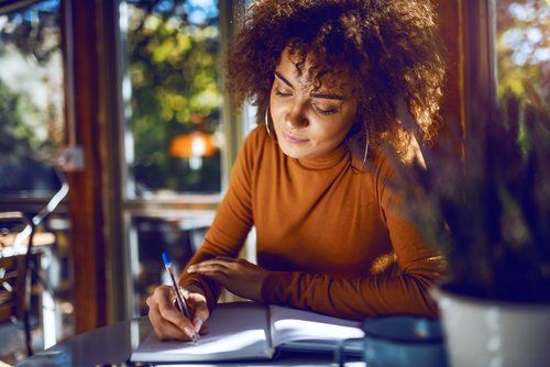 A woman with curly hair sits at a table, writing in a notebook while working in a bright café.