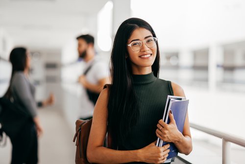 Eine junge Frau mit langen, schwarzen Haaren und Brille hält Bücher in der Hand und lächelt, während sie in einem modernen Lernumfeld steht.