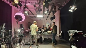 Two people are standing at a table with camera and lighting equipment, while a group watches in the background.