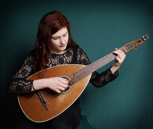 A woman with long, red hair is playing a mandolin while sitting against a green background. She is wearing a black top with lace detailing.