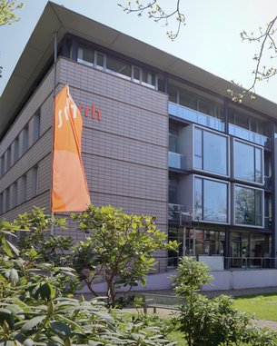 The building with large windows and a modern design features the logo "sitch" on its facade. In front of the building, there are green plants and an orange flag visible.