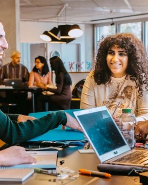 A group of five people is sitting at a table, engaging in lively discussion and smiling. On the table are notebooks, a laptop, and various writing utensils. In the background, more people can be seen.