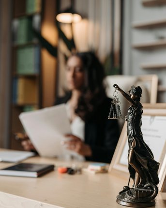 A woman is sitting at a desk, holding documents in her hand. In the foreground stands a statue of Justice holding a scale. The background features a modern office environment with shelves and plants.