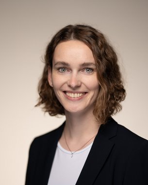 A smiling woman with curly brown hair is wearing a black blazer and a white blouse. The background is neutral and bright.