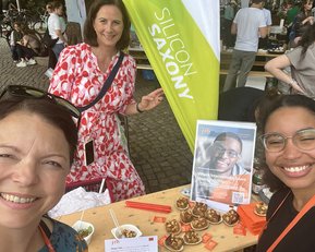 Drei Frauen posieren lächelnd vor einem Stand mit Speisen und einem Plakat. Im Hintergrund sind weitere Personen und Stände zu sehen.