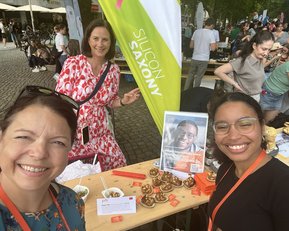 Drei Frauen posieren lächelnd vor einem Stand mit Speisen und einem Plakat. Im Hintergrund sind weitere Personen und Stände zu sehen.