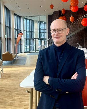 A man with glasses and a black turtleneck stands in a modern room with large windows and orange lamps.