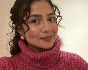 Eine Person mit lockigem Haar trägt einen rosa Rollkragenpullover und lächelt in die Kamera. Der Hintergrund ist neutral.