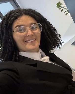 A person with curly hair and glasses is wearing a suit and posing in front of a desk with a computer and keyboard.