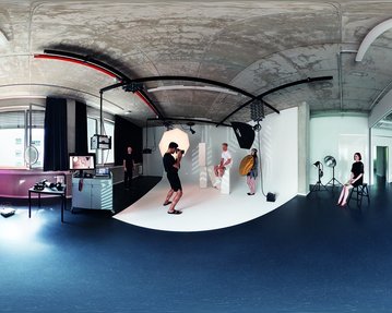 Ein Studio mit einem runden Fotohintergrund, einem Fotografen, der ein Model ablichtet. Im Hintergrund sind weitere Personen und Ausrüstungen sichtbar. Die Wände sind hell, und der Boden ist dunkel.