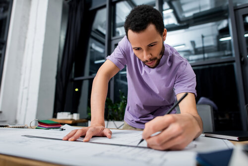 A man is focused on drawing on a large sheet of paper in a modern office. Tools and materials are scattered around him.