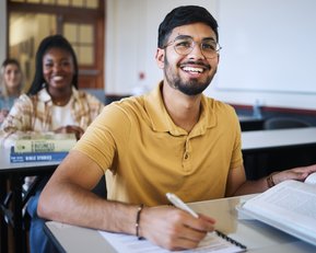 Ein junger Mann mit Brille sitzt an einem Schreibtisch in einem Klassenzimmer, lächelt und hält ein Buch. Im Hintergrund sind zwei weitere Studierende zu sehen, die ebenfalls an ihren Schreibtischen sitzen.
