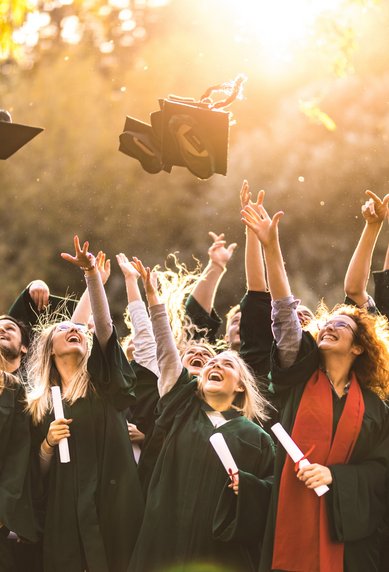Graduates in academic attire celebrate their graduation by throwing their caps into the air. Some are holding their diplomas in their hands. The scene is bathed in sunlight, creating a festive atmosphere.
