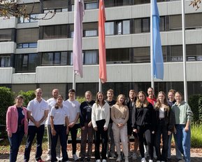 Gruppe von 18 Personen steht vor einem Gebäude, umgeben von Fahnen. Die Teilnehmer tragen verschiedene Freizeitkleidung und lächeln.