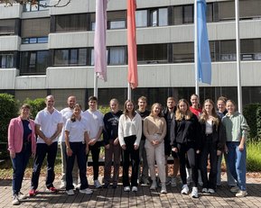 Gruppe von 18 Personen steht vor einem Gebäude, umgeben von Fahnen. Die Teilnehmer tragen verschiedene Freizeitkleidung und lächeln.