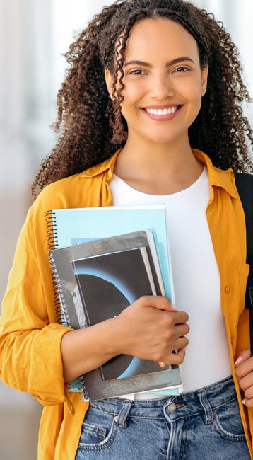 Eine junge Frau mit lockigem Haar trägt eine orangefarbene Jacke und hält mehrere Bücher in den Händen. Sie hat einen schwarzen Rucksack über der Schulter und steht in einem modernen Innenraum.