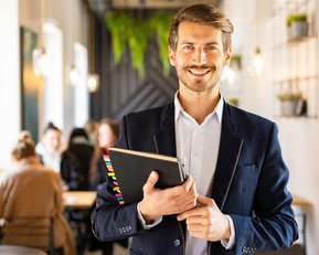 Ein Mann in einem blauen Blazer hält ein Notizbuch und lächelt. Im Hintergrund sind unscharfe Personen in einem Café zu sehen. Die Atmosphäre wirkt freundlich und einladend.