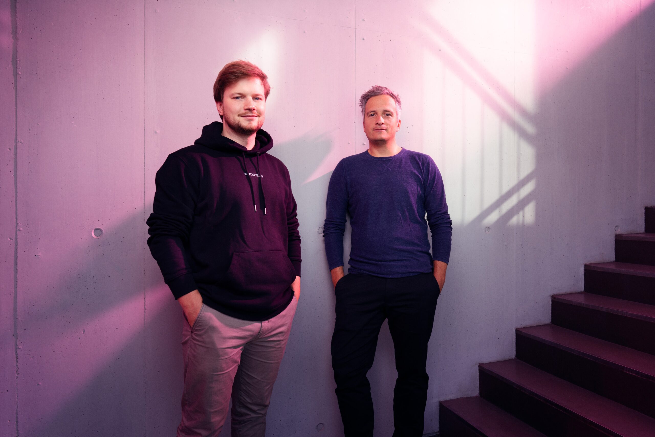 Two men stand side by side in front of a wall with soft light reflections. The man on the left is wearing a black hoodie, while the man on the right is in a blue top. In the background, stair steps can be seen.