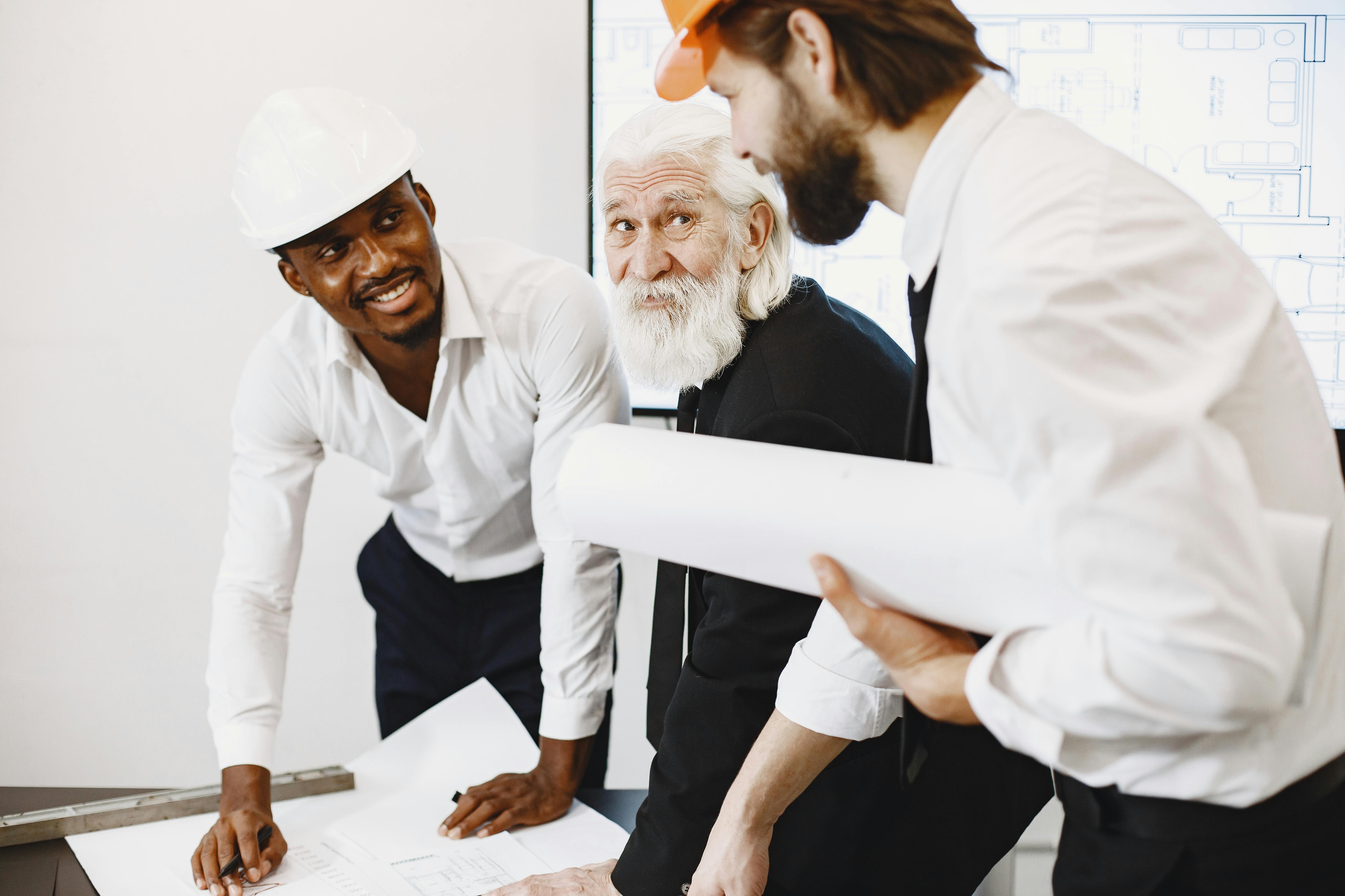 Drei Männer in einem Büro besprechen Baupläne. Einer trägt einen weißen Helm, der andere einen orangefarbenen. Der ältere Mann schaut interessiert.