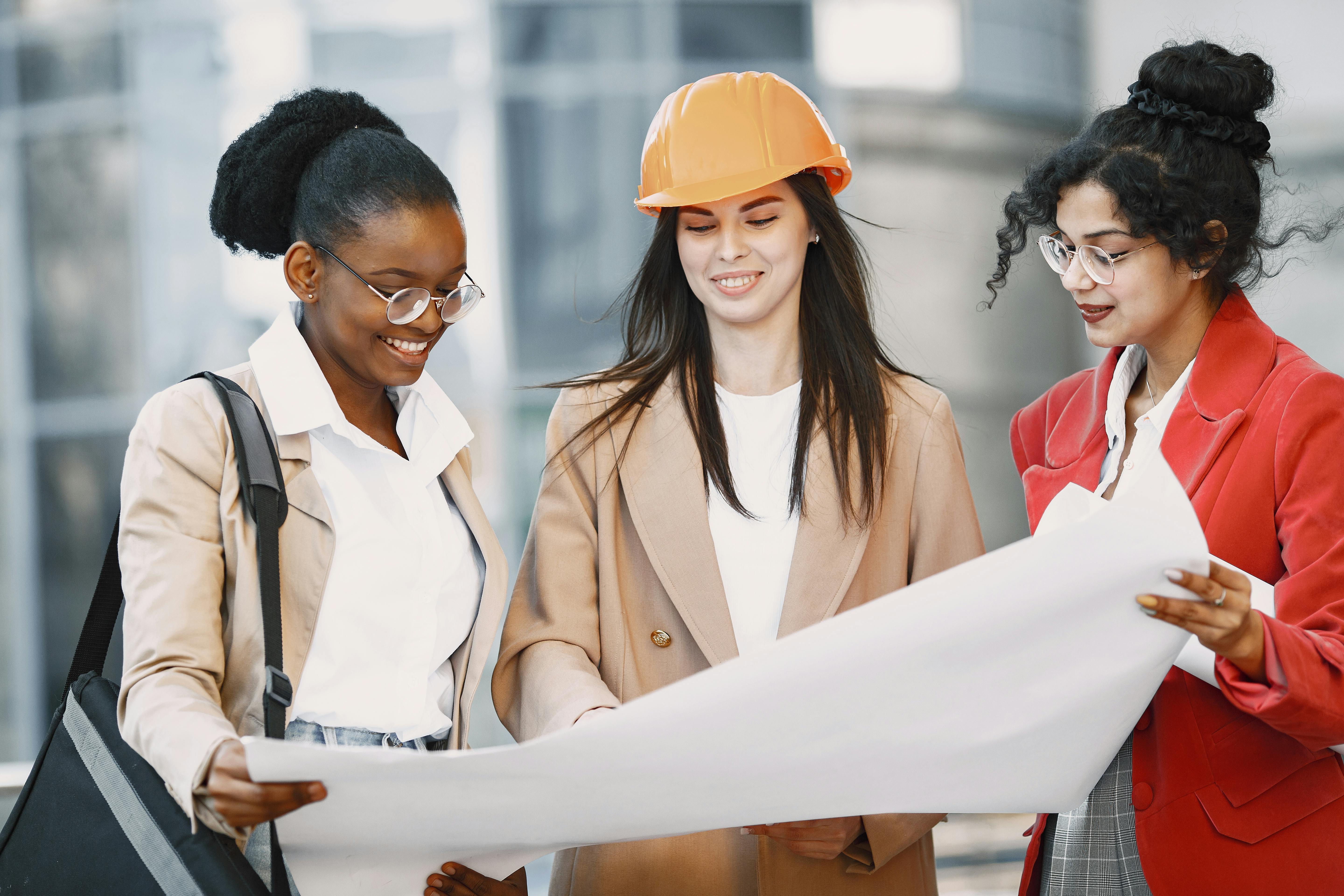 Drei Frauen betrachten gemeinsam Baupläne, eine trägt einen orangefarbenen Helm. Sie stehen in einem modernen, urbanen Umfeld.