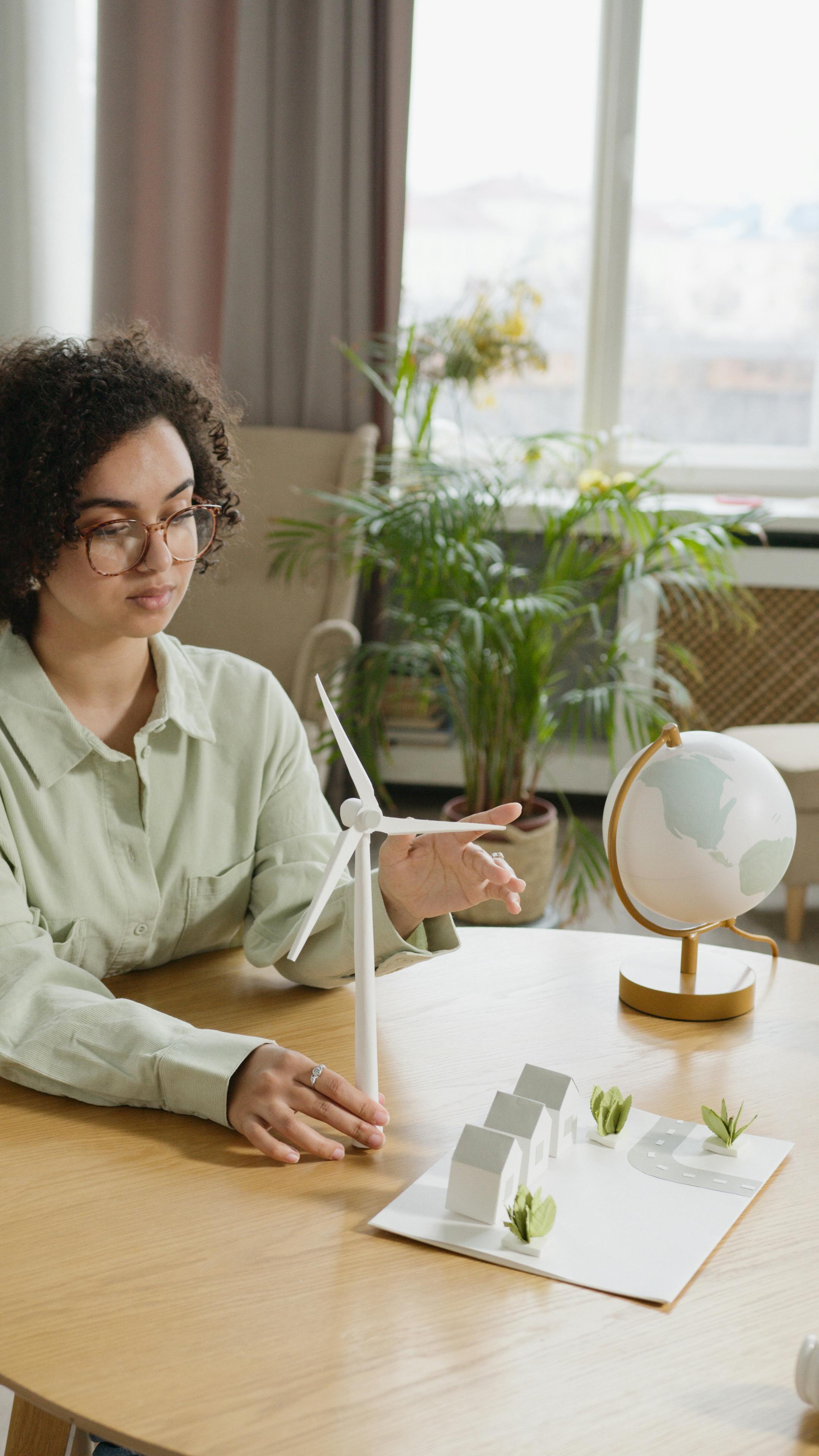Eine Frau mit lockigem Haar und Brille zeigt auf ein Modell eines Windrads und ein Miniaturdorf auf einem Tisch. Ein Globus steht daneben.